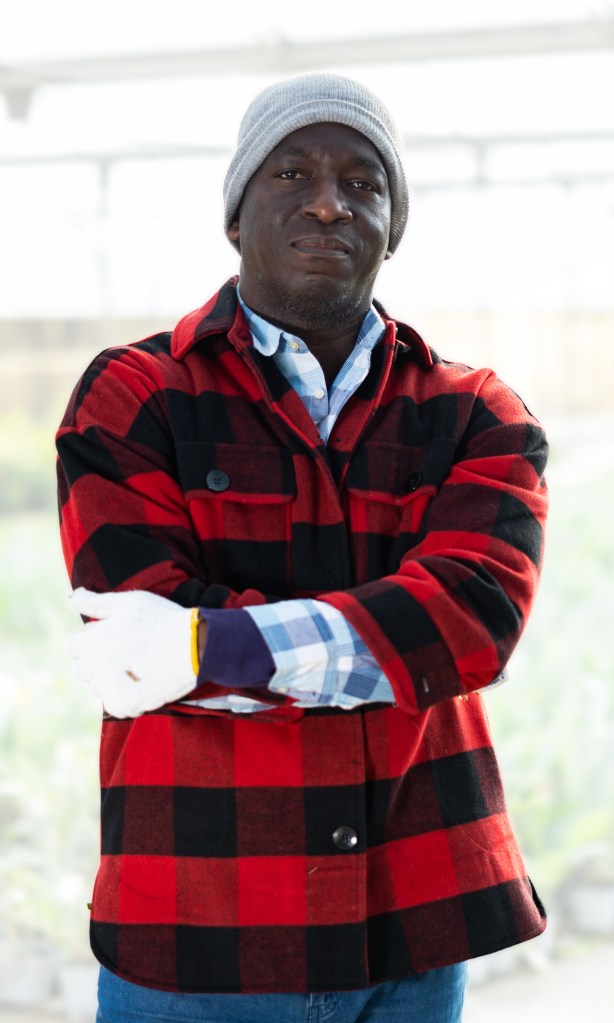 Derek standing in greenhouse with arms folded looking serious