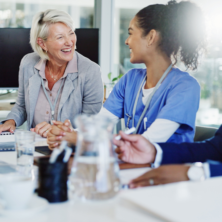 Woman in business suit laughing at a table with female nurse wearing blue scrubs.