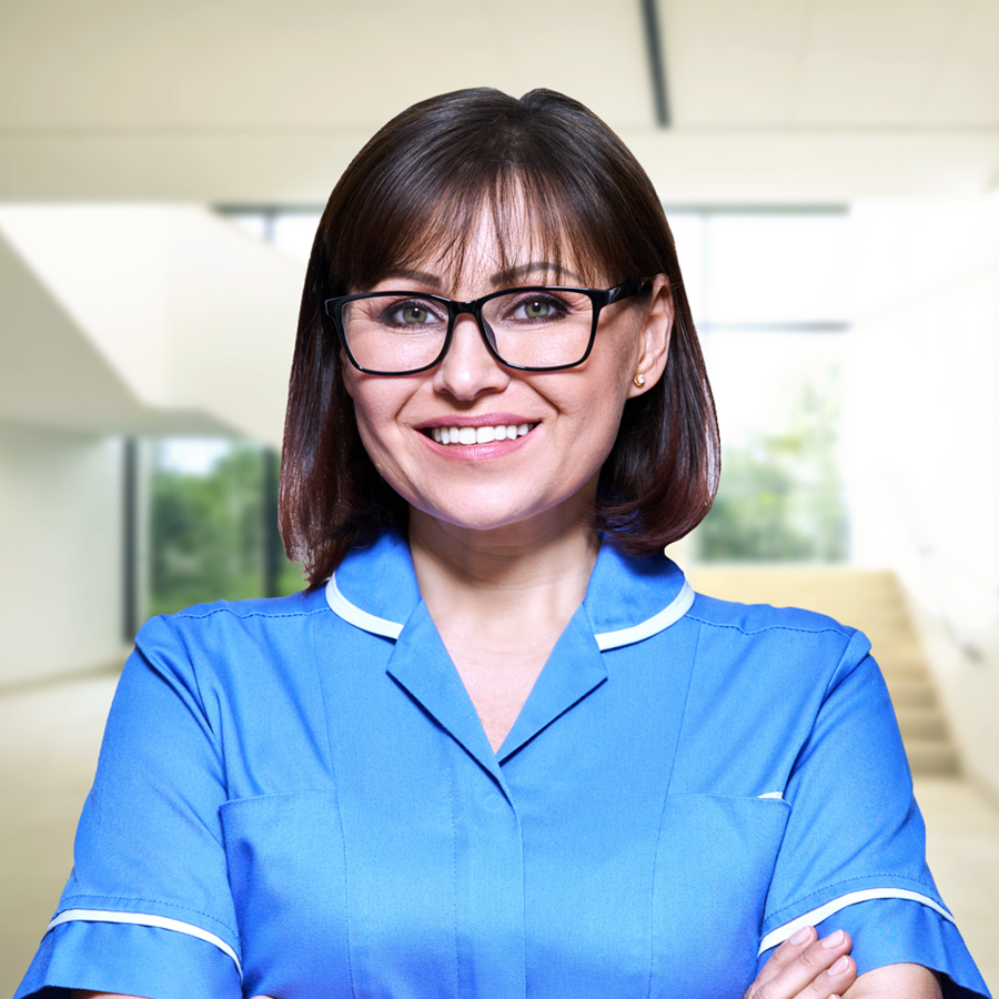 Female nurse wearing blue uniform and smiling, standing in a modern building.
